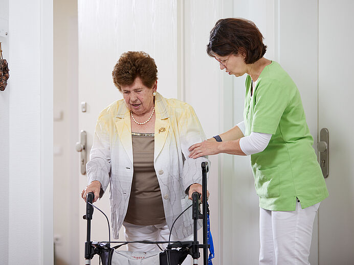 Ambulanter Pflegedienst: Dame hilft älterer Dame mit Rollator beim Betreten einer Wohnung.